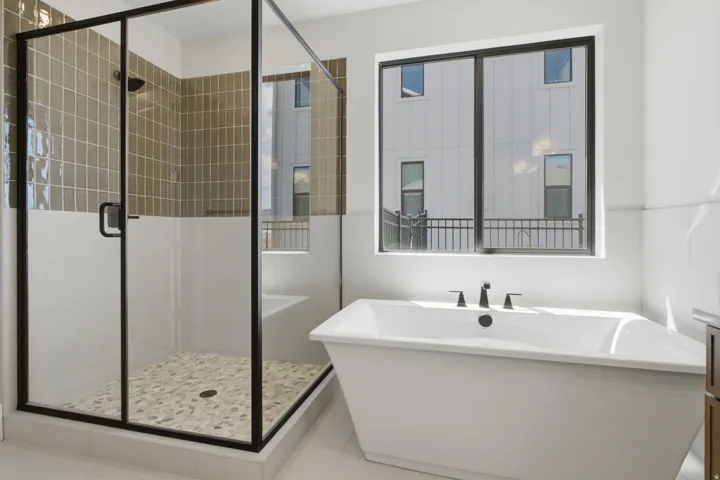 Freestanding soaking tub with a black faucet, glass-enclosed shower with a black frame, textured tile shower surround, pebble tile shower floor, and a large window