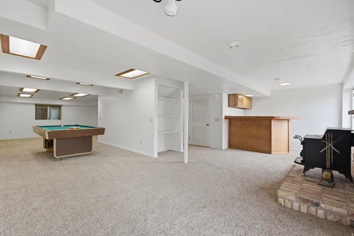 Carpeted recreational area featuring a built-in bar with wood-finish cabinetry, a brick fireplace with a wood-burning stove, built-in shelving, and recessed lighting