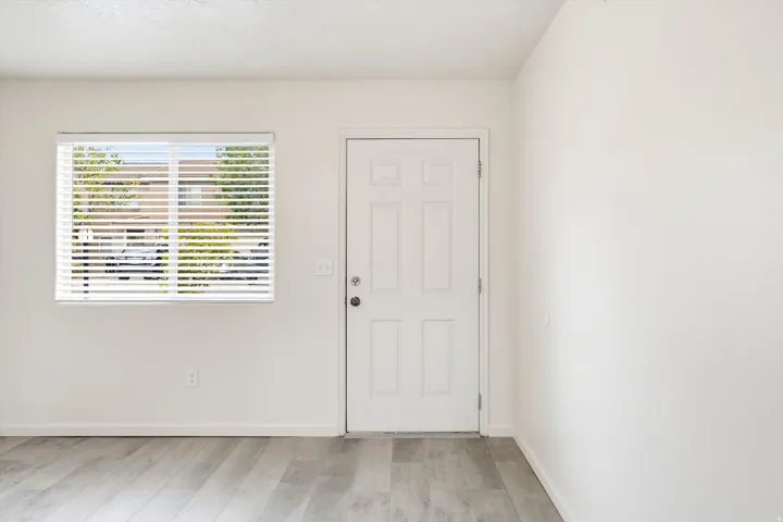 White interior door with paneled design, accompanied by a window with horizontal blinds, set against light-toned walls and wood-finish flooring