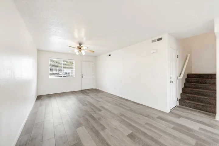 Spacious room featuring wood-finish flooring, a ceiling fan with integrated lighting, a window with blinds, and a staircase with carpeted treads