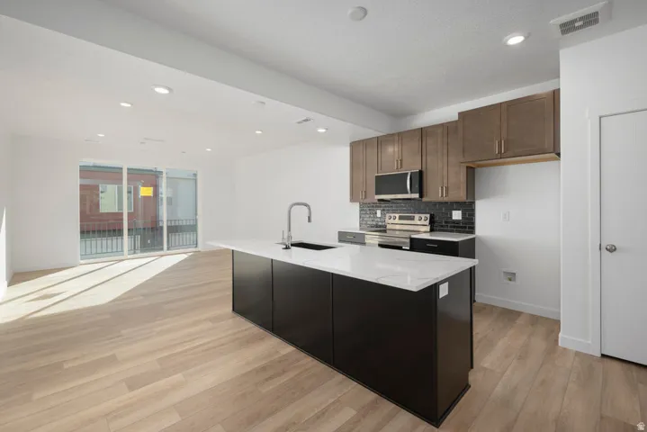 Modern kitchen and open living space featuring custom cabinetry in warm wood tones, a striking black-finished island with white quartz countertop, stainless steel appliances, and wide-plank wood-finish flooring throughout