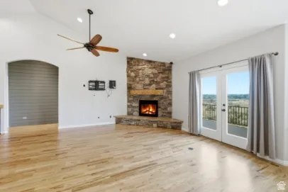 Vaulted ceiling with a ceiling fan, stone-clad fireplace with a wood mantle, wood-finish flooring, and double patio doors