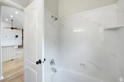 Bathtub with a built-in shower, white tile surround with subtle veining, and a corner shelf