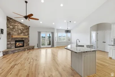 Vaulted ceilings with recessed lighting, stone-clad fireplace with wood mantel, light wood-finish flooring, white double French doors, and a kitchen island with a granite countertop
