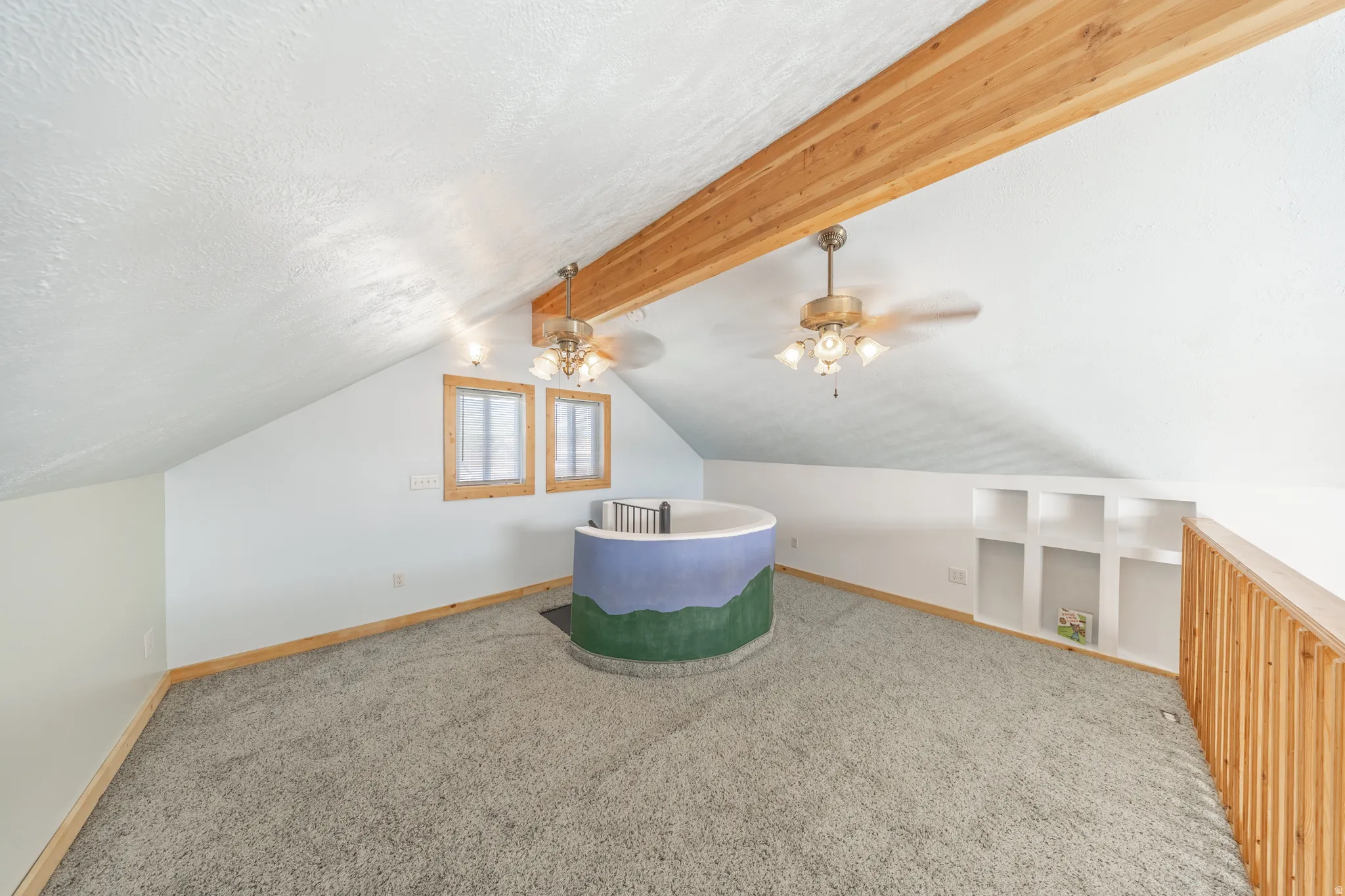 Vaulted ceiling with exposed wood beams, two ceiling fans with light kits, two windows with wood trim, built-in shelving, and carpeted flooring