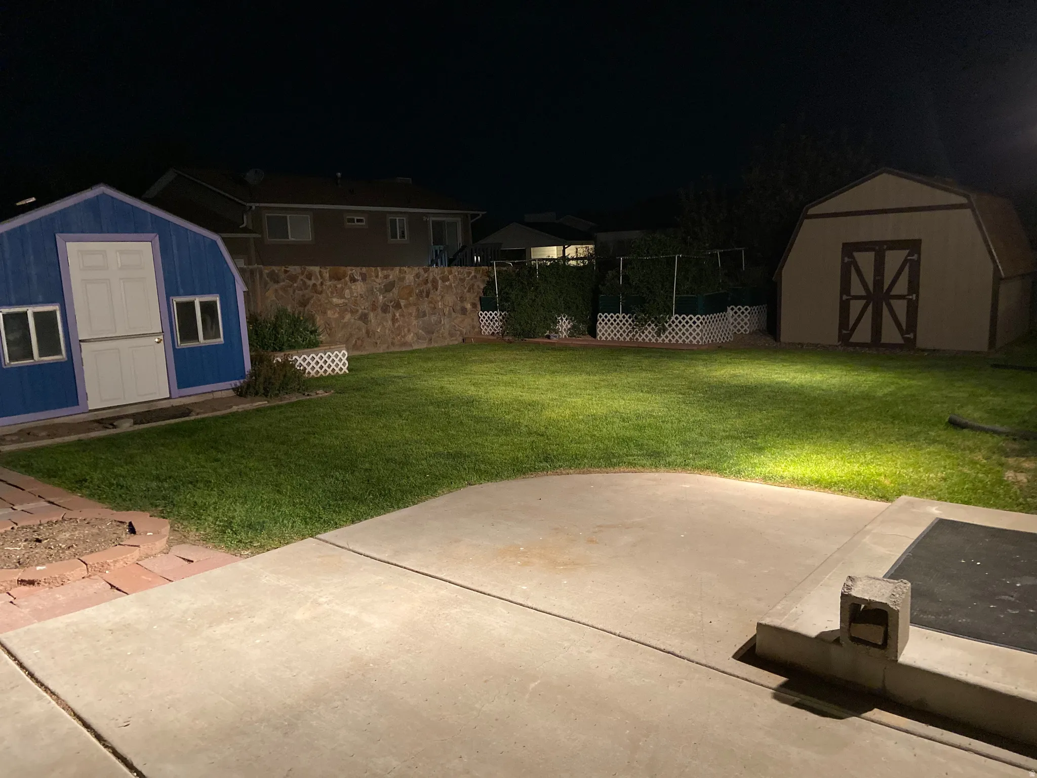 Expansive backyard featuring a concrete patio, lush green lawn, full-height stone retaining wall, and two storage sheds