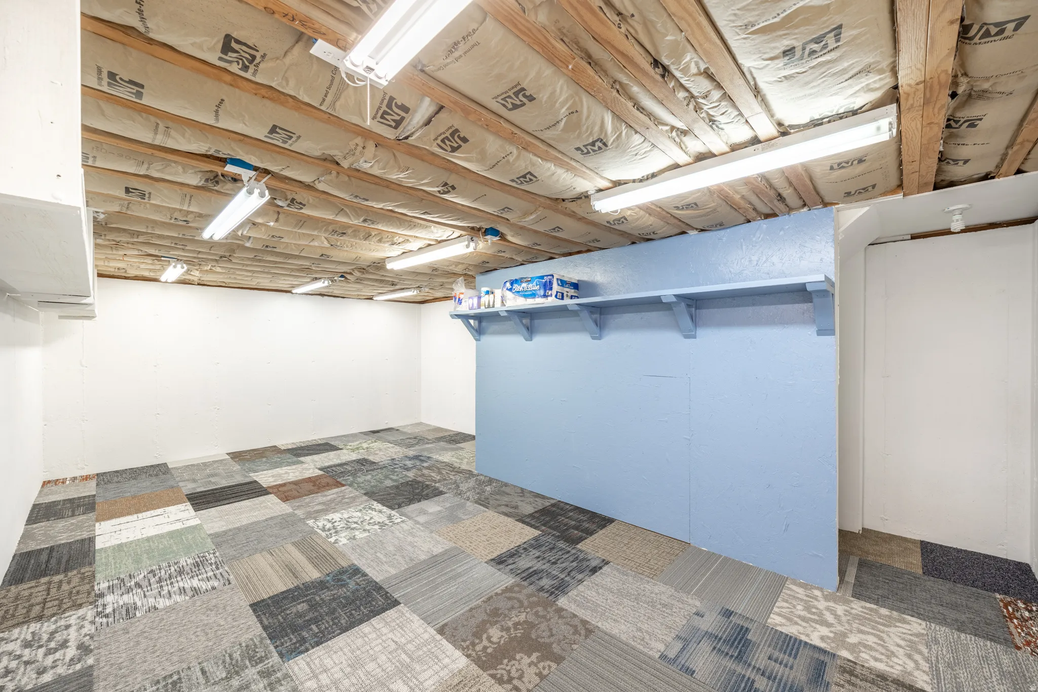 Exposed ceiling joists with insulation, fluorescent lighting fixtures, light blue accent wall, built-in shelving, and multi-patterned carpet tile flooring