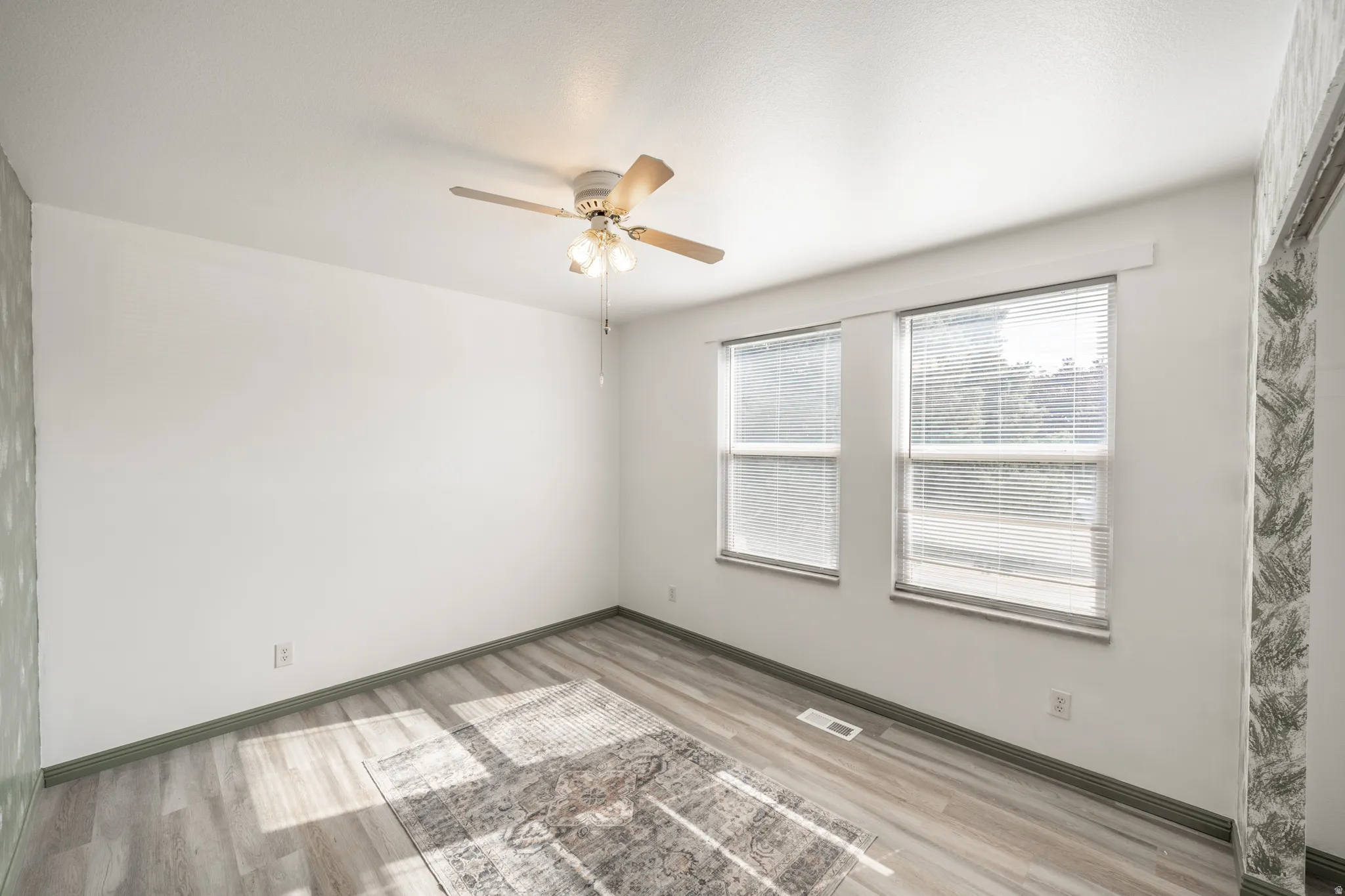 Wood-finish flooring with a light gray tone, two windows with blinds, a ceiling fan with light fixture, and white painted walls