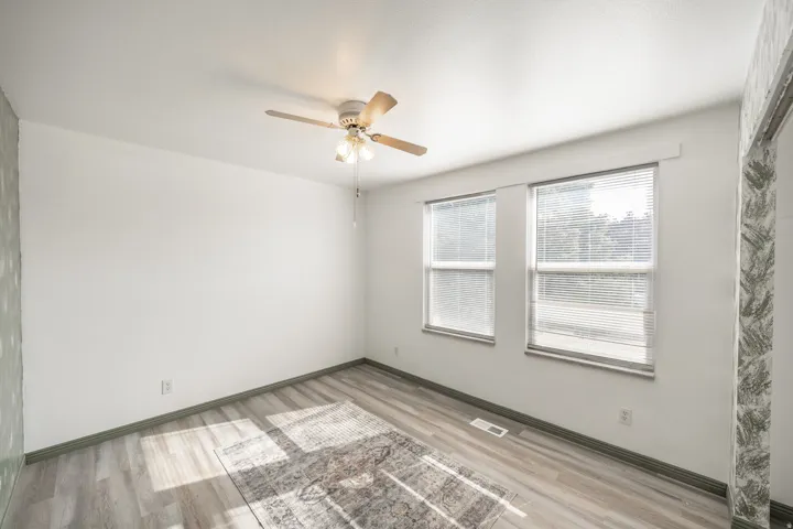 Wood-finish flooring with a light gray tone, two windows with blinds, a ceiling fan with light fixture, and white painted walls