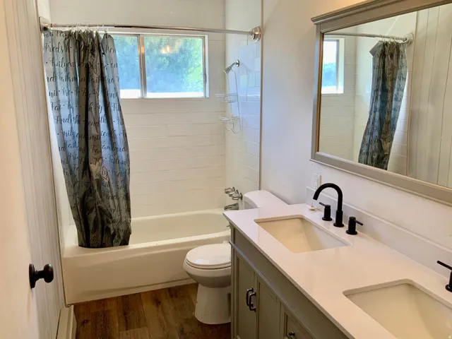 Double vanity with matte black faucets, wood-finish flooring, white subway tile shower surround, and a framed mirror