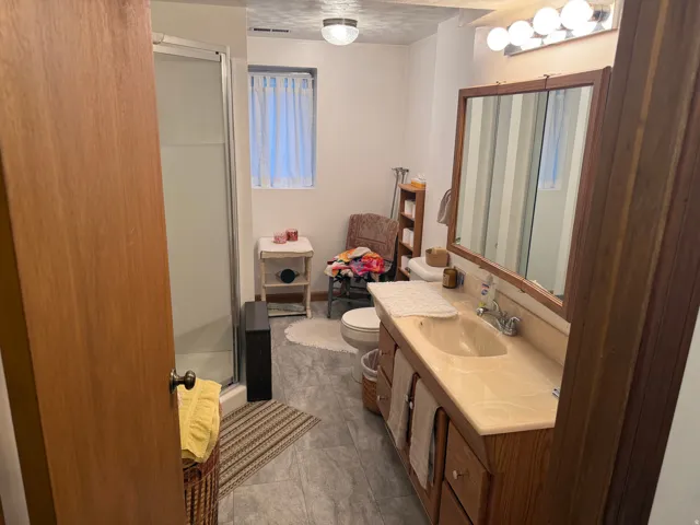 Wood-finish vanity with an integrated sink and light-colored countertop, framed mirror with overhead lighting, tile flooring, and a walk-in shower with a frosted glass enclosure