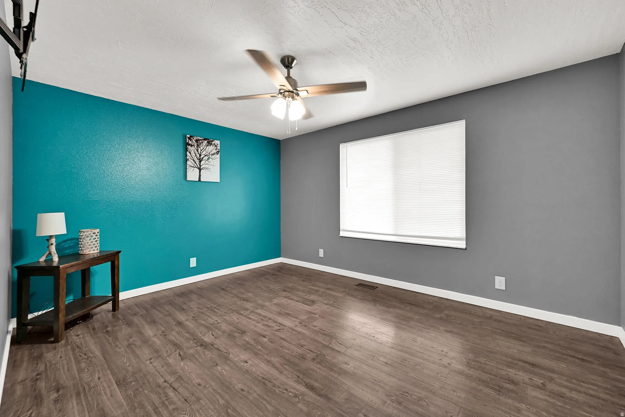 Wood-finish flooring, ceiling fan with integrated lighting, window with privacy blind, and accent wall