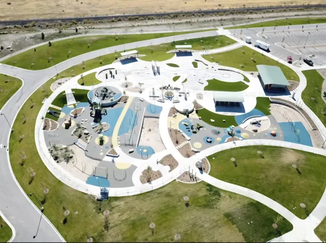 Expansive green spaces with paved walking paths, multiple shaded structures, and a large recreational play area featuring diverse activity zones