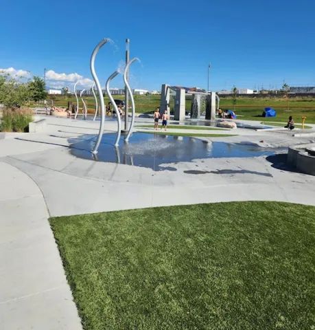 Modern waterfront park featuring sculptural stainless steel water features, shallow reflecting pool with integrated water jets, and expansive concrete hardscape