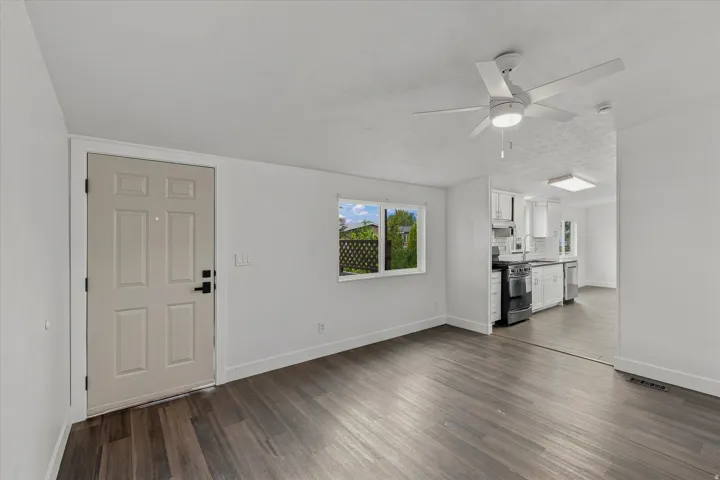 White interior walls, wood-finish flooring, white ceiling fan with integrated lighting, white paneled entry door with black hardware, and a window with exterior landscaping