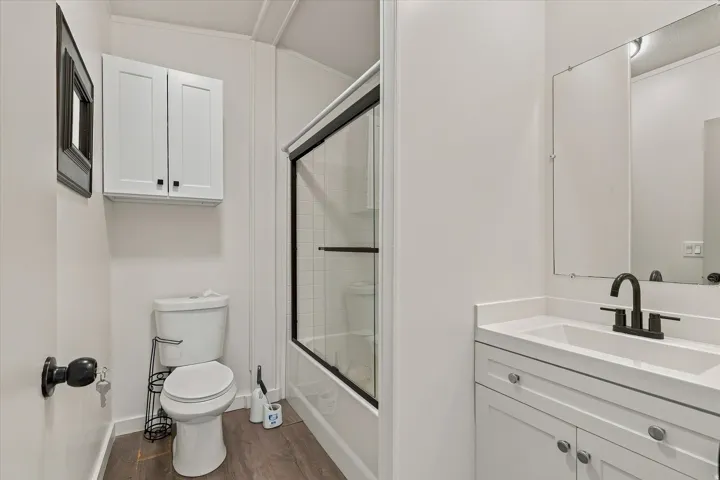 Modern bathroom featuring a white vanity with an undermount sink and matte black faucet, quartz countertop, white cabinetry with hardware accents, large framed mirror, wood-finish flooring, frameless glass shower enclosure, and white subway tile walls thr