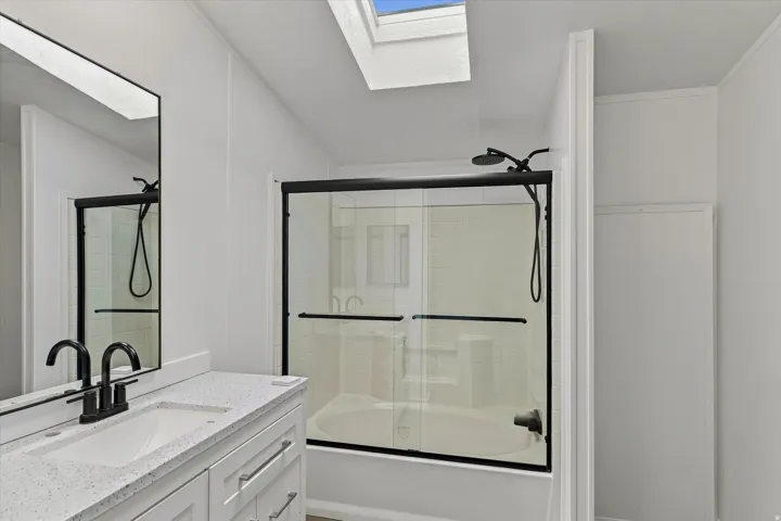 Built-in bathtub with glass sliding doors and matte black fixtures, integrated vanity with a speckled countertop and undermount sink, matte black faucet, and rectangular wall mirror