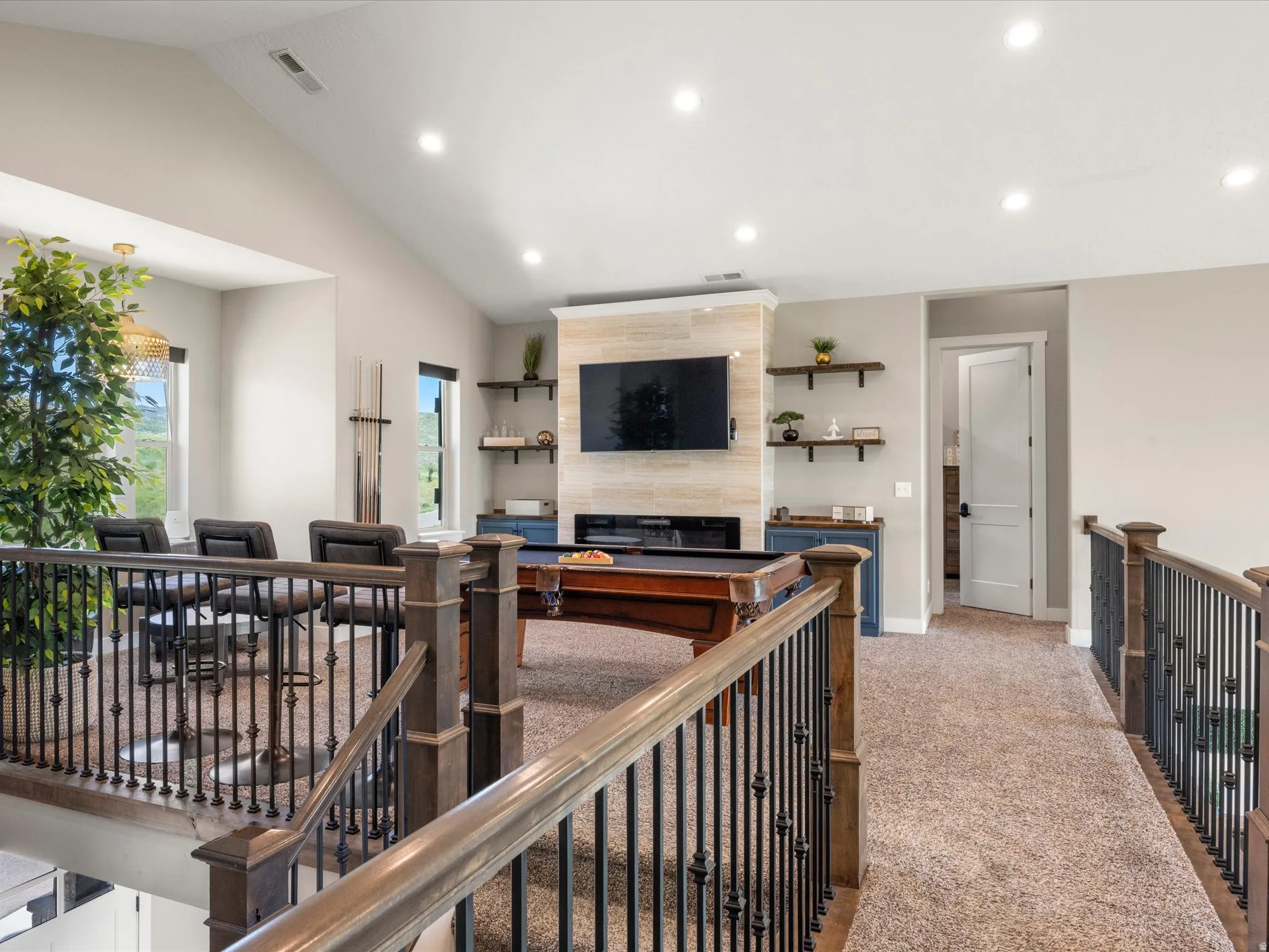 Carpeted loft area featuring a vaulted ceiling with recessed lighting, wood-finish and wrought iron railings, a wall-mounted television above a tiled accent wall, and built-in shelving