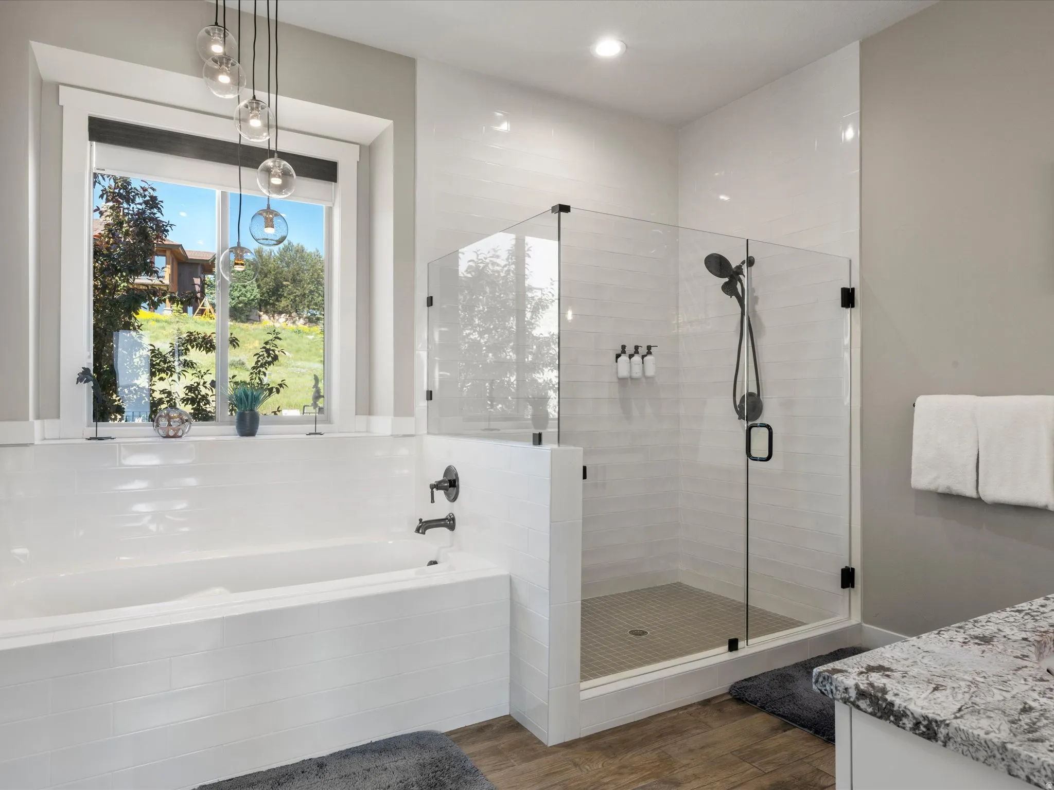 Freestanding soaking tub with matte black fixtures, glass-enclosed shower with subway tile, wood-finish flooring, and a window with pendant lighting