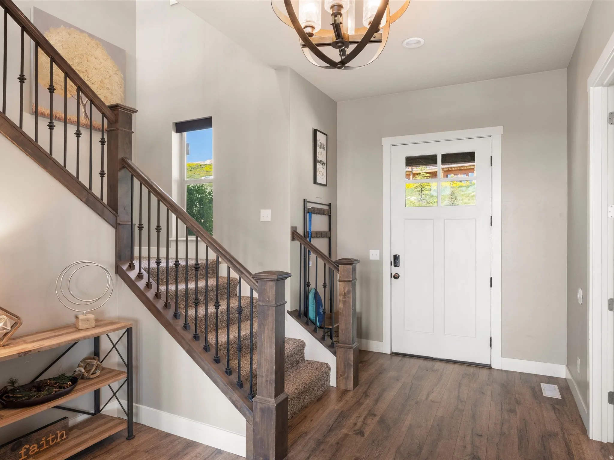 Wood-finish flooring, a white paneled entry door with glass inserts, a wood staircase with carpeted treads and wrought iron balusters, and a modern chandelier