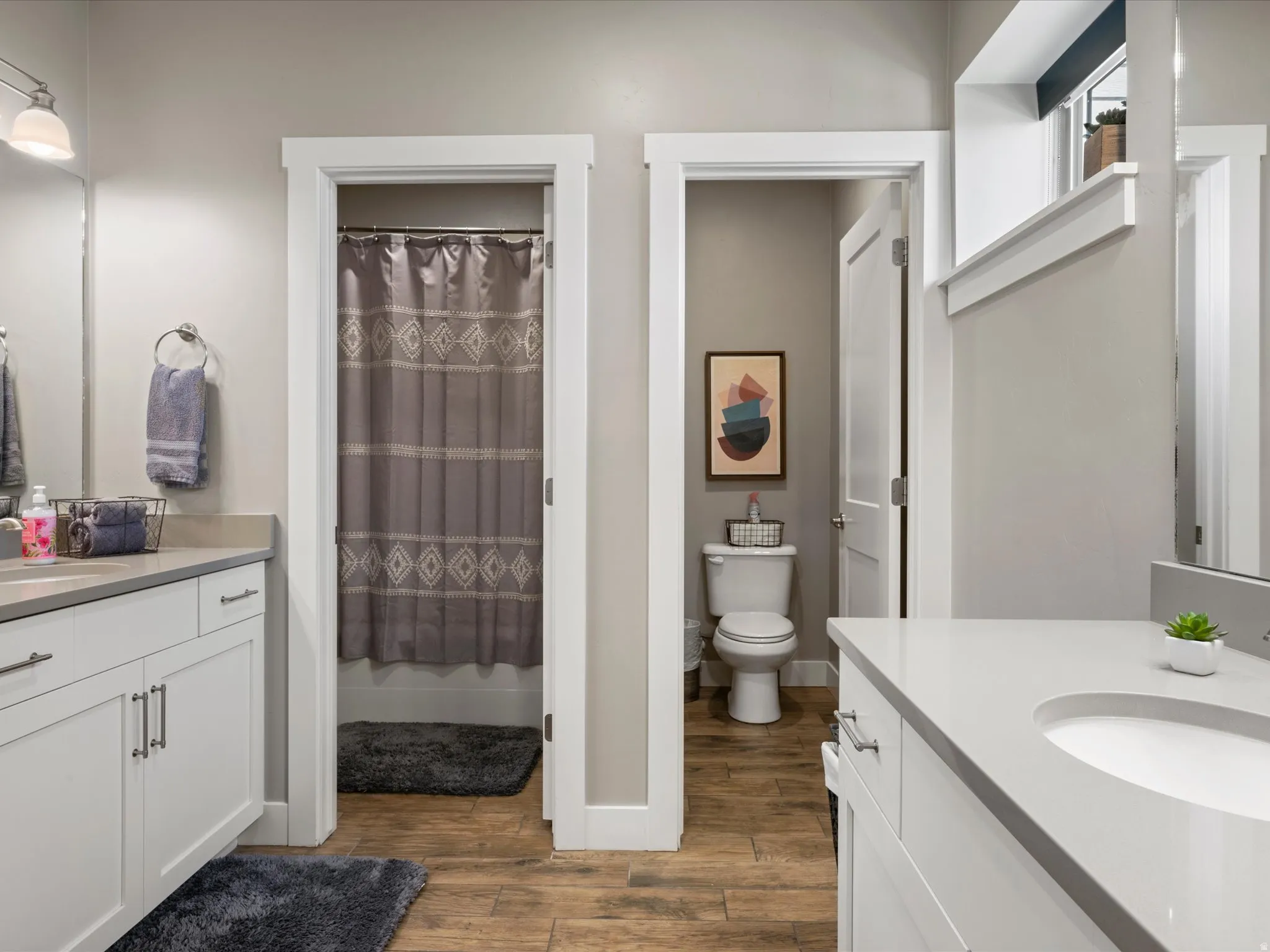 Dual white vanities with integrated sinks, light-toned countertops, and wood-finish flooring