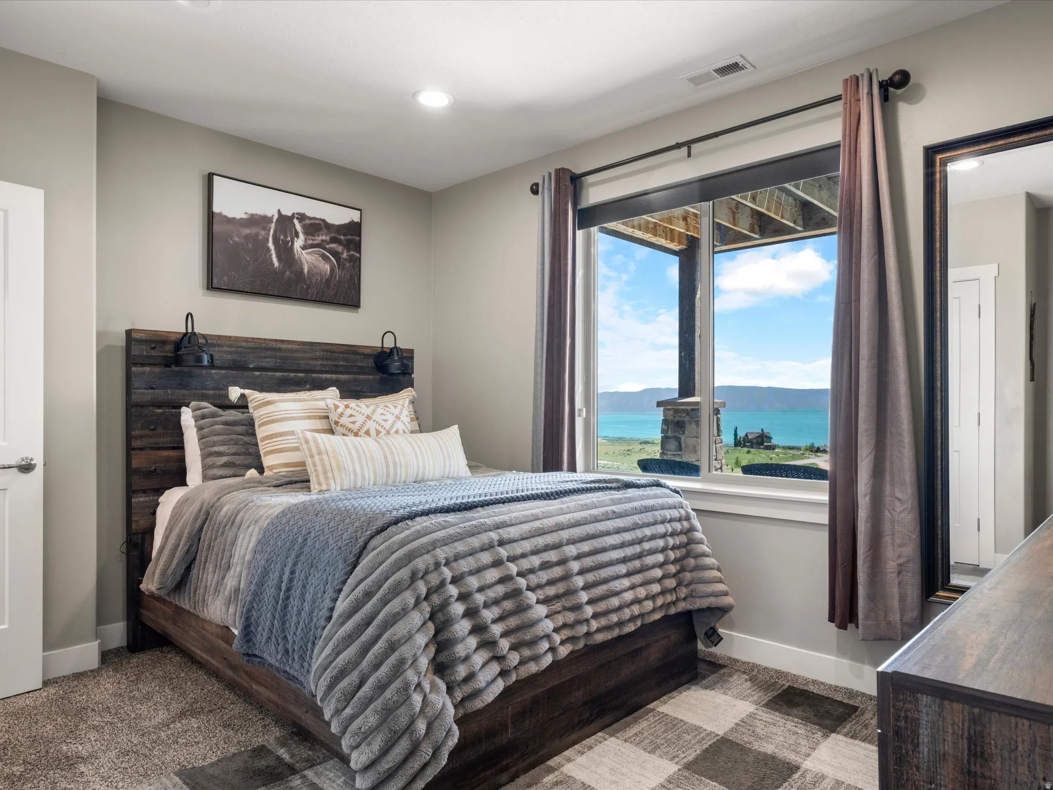 Recessed ceiling lighting, carpeted flooring with area rug, wood-finish headboard with integrated sconce lighting, window with mountain and water scenery