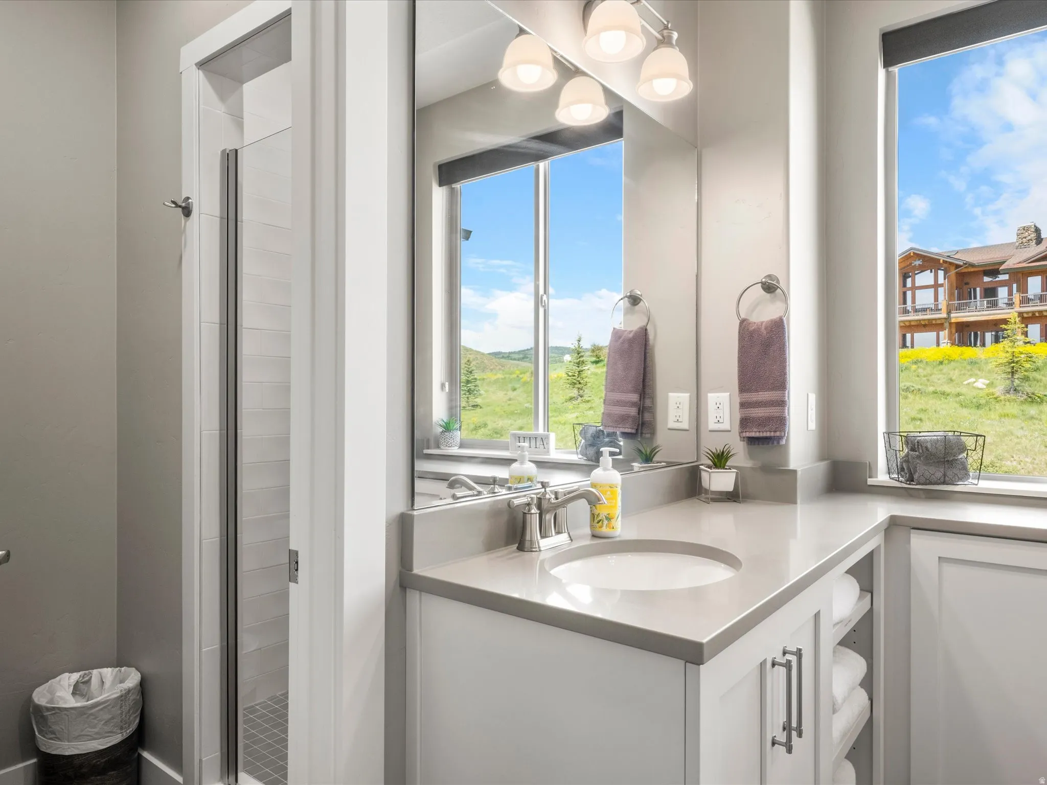 Walk-in shower with glass enclosure, single vanity with integrated sink, stone countertop, built-in storage shelving, and large windows
