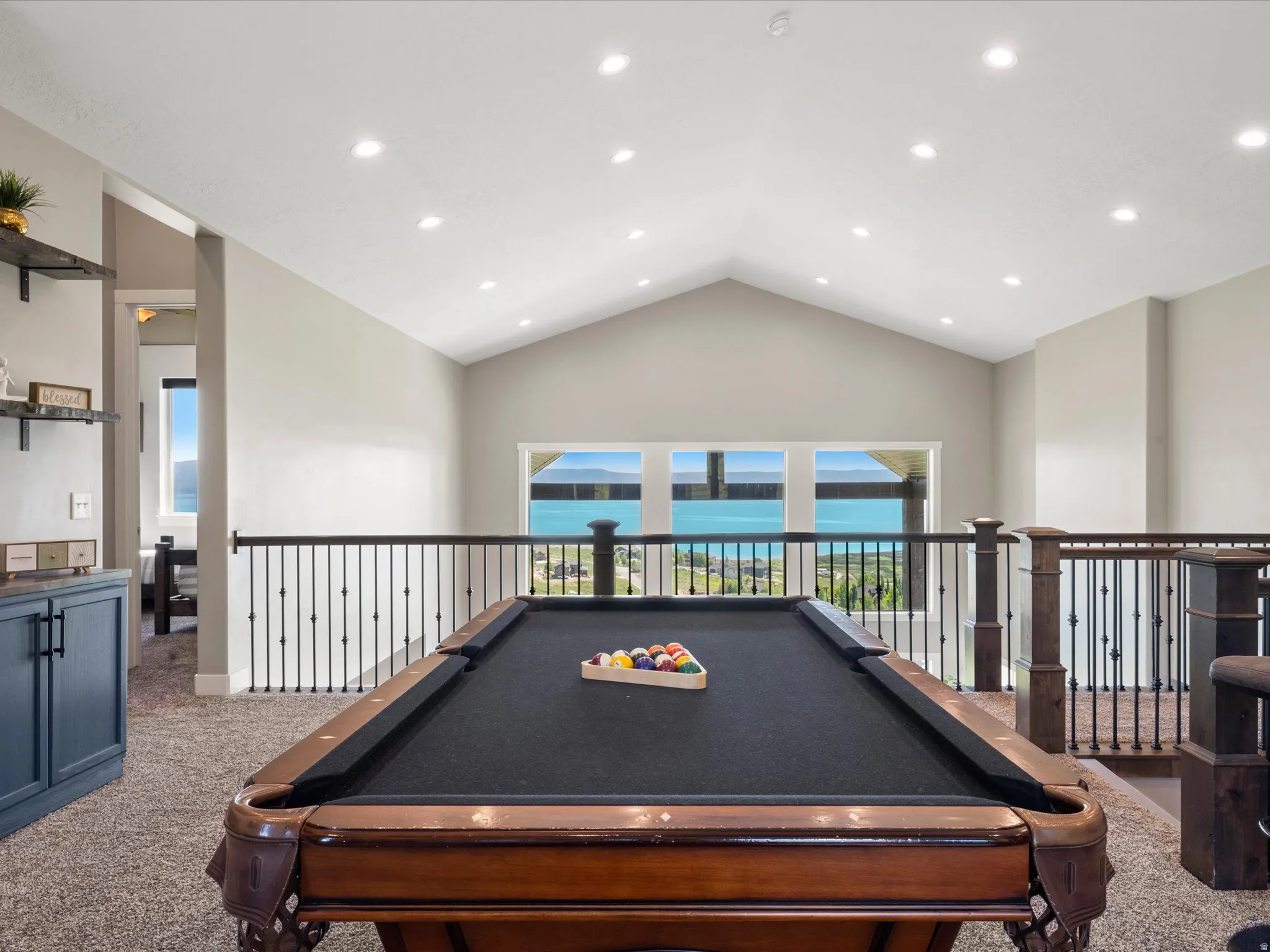 Vaulted ceiling with recessed lighting, large windows, dark wood-finish railing with black metal spindles, built-in shelving, and a built-in cabinet with dark blue panel doors