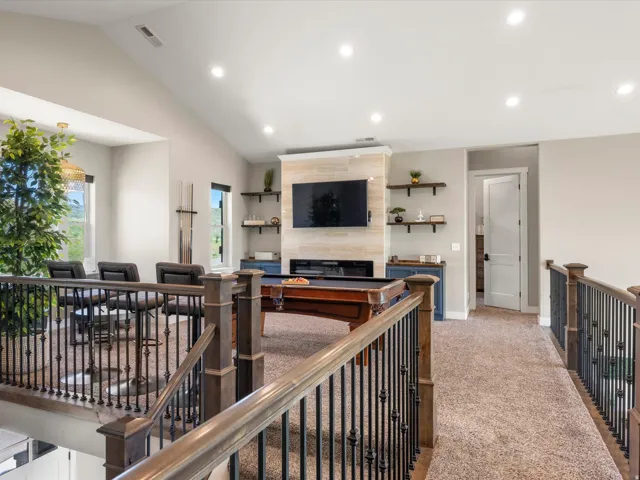 Carpeted loft area featuring a vaulted ceiling with recessed lighting, wood-finish and wrought iron railings, a wall-mounted television above a tiled accent wall, and built-in shelving