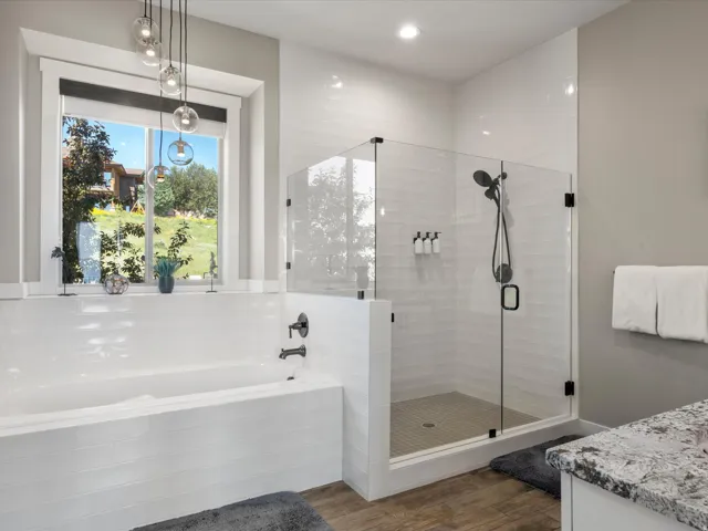 Freestanding soaking tub with matte black fixtures, glass-enclosed shower with subway tile, wood-finish flooring, and a window with pendant lighting