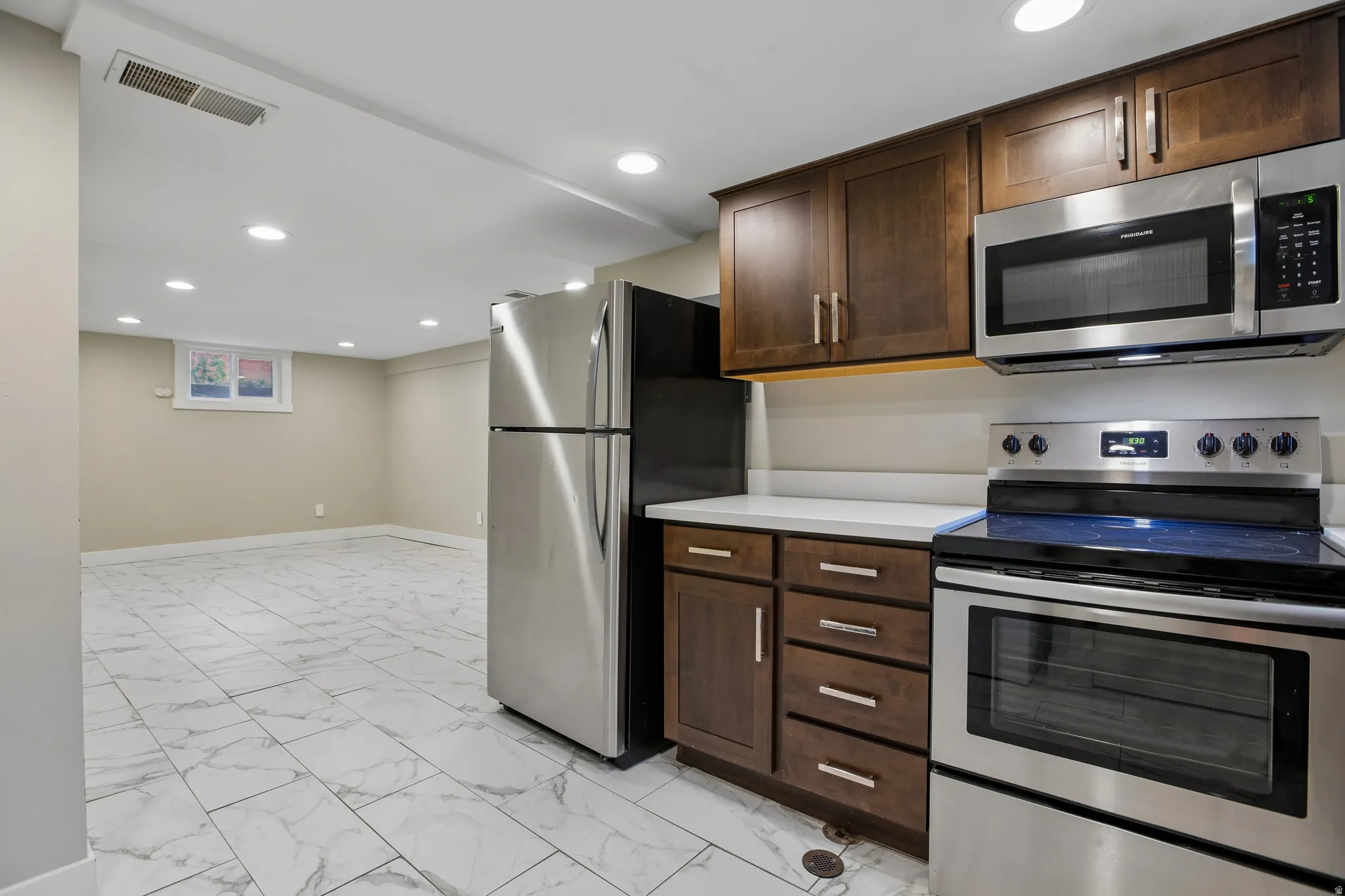 Stainless steel appliance suite, dark wood-finish cabinetry, light-toned countertops, recessed lighting, and large format tile flooring