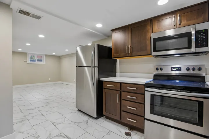 Stainless steel appliance suite, dark wood-finish cabinetry, light-toned countertops, recessed lighting, and large format tile flooring