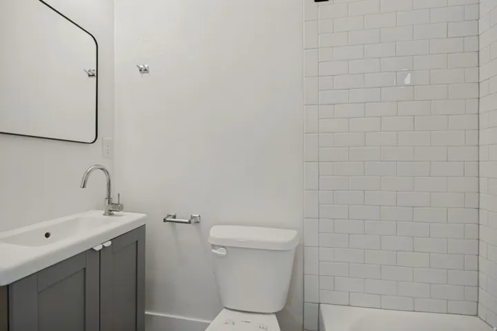 Wall-mounted mirror, vanity with integrated sink, chrome faucet, white subway tile shower surround, and a white toilet