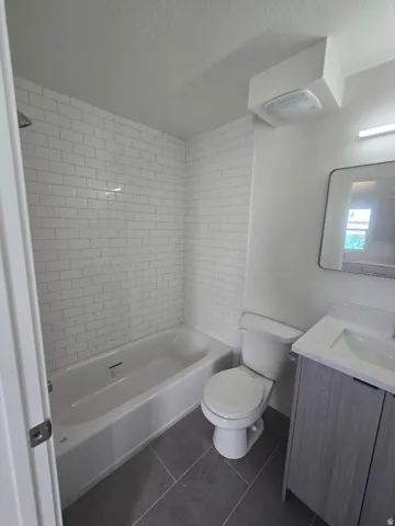 White subway tile shower surround, integrated bathtub, gray rectangular floor tiles, modern vanity with a white countertop and integrated sink, and an overhead light fixture
