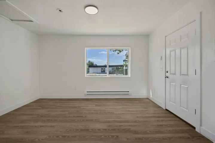 Spacious room featuring wood-finish flooring, large window with natural light, baseboard heating unit, and white six-panel closet doors