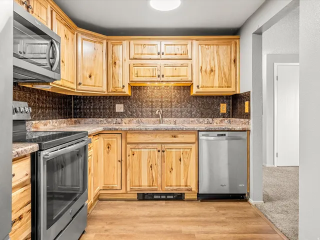 Kitchen featuring wood-finish cabinetry, stainless steel appliances, a textured dark backsplash, and wood-finish flooring