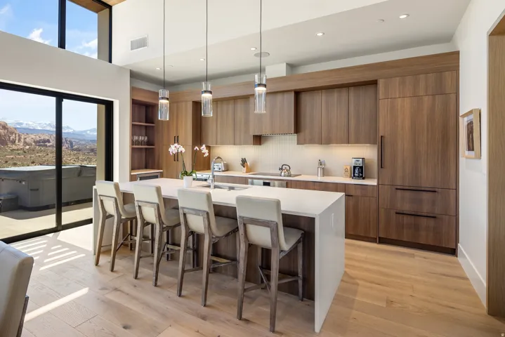Modern kitchen featuring light wood-finish flooring, a large center island with seating, dark wood-finish cabinetry, built-in panel-ready appliances, and a full wall of glass sliding doors opening to a patio with a distant mountain backdrop