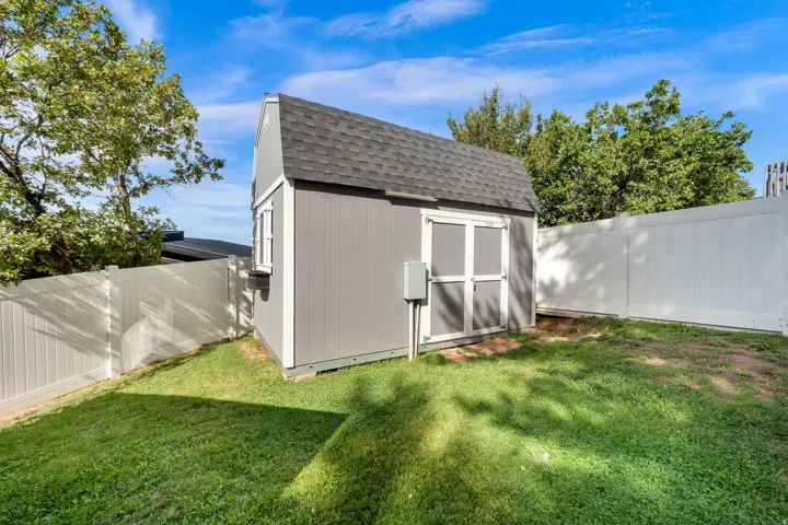 Gable-roofed storage shed with double doors and window, surrounded by a white privacy fence and a grassy area
