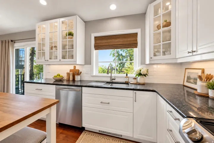 White cabinetry with glass-front uppers, stainless steel sink, black countertops, white subway tile backsplash, and stainless steel dishwasher