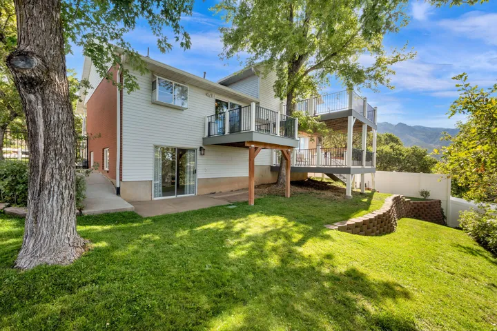 Rear exterior featuring a two-story deck with metal railings, a ground-level patio, a block retaining wall, and mature trees