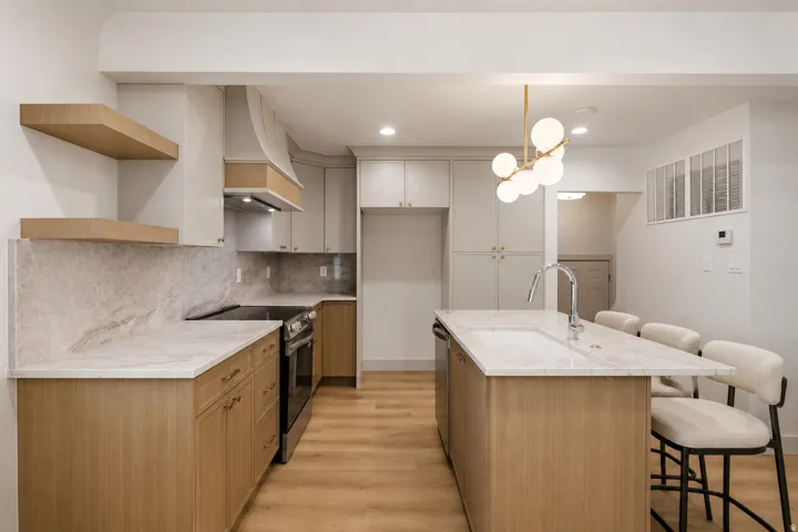 Kitchen featuring a large island with an integrated sink, light wood-finish cabinetry, stone countertops and backsplash, and a modern chandelier