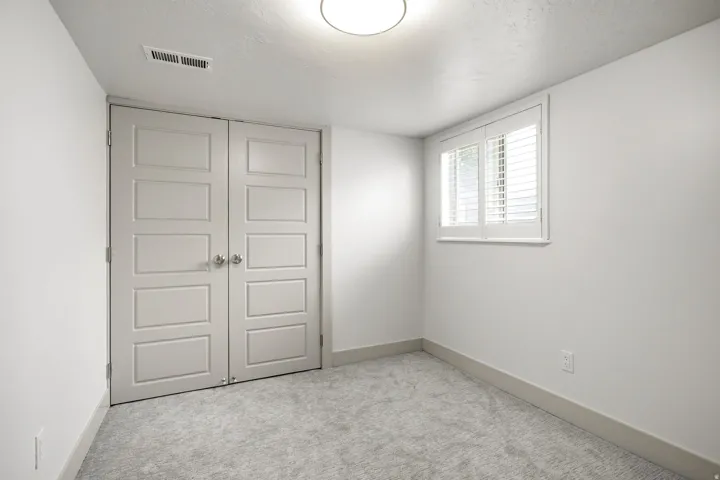 White walls, light-colored carpeting, a window with plantation shutters, and a flush-mount ceiling light fixture