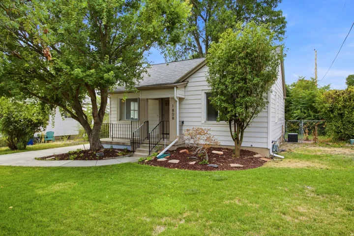 White siding exterior with a dark shingle roof, mature trees, concrete driveway, and front porch with black railings