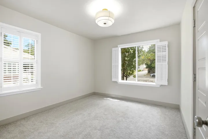 Light gray carpet flooring, two windows with white plantation shutters, white paneled interior door, and a flush-mount ceiling light fixture