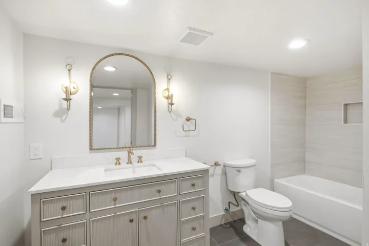 Vanity with gold-toned hardware, arched mirror with gold-toned frame, wall-mounted sconces, white countertop, and tiled shower surround with built-in niche