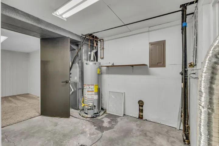 Utility area with concrete flooring, water heater, visible piping, and access to a separate carpeted room