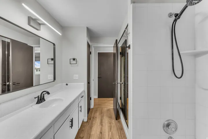Bathroom featuring a single vanity with a white countertop, matte black fixtures, a rectangular mirror, wood-finish flooring, and a tiled shower with a built-in shelf