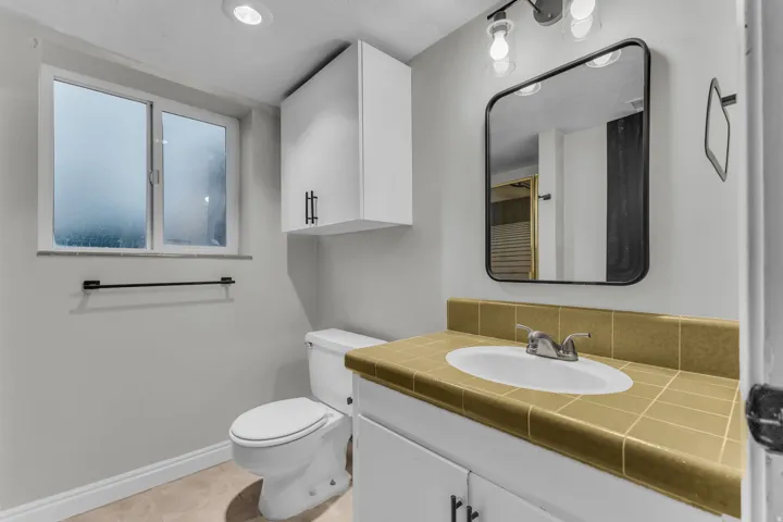 Vanity with tiled countertop and integrated sink, white cabinetry with dark hardware, and a wall-mounted mirror with an overhead light fixture