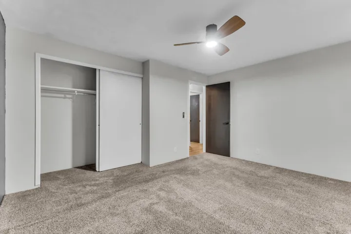 Neutral-toned carpeting, white walls, built-in closet with sliding doors, ceiling fan with light fixture, and dark-paneled interior door