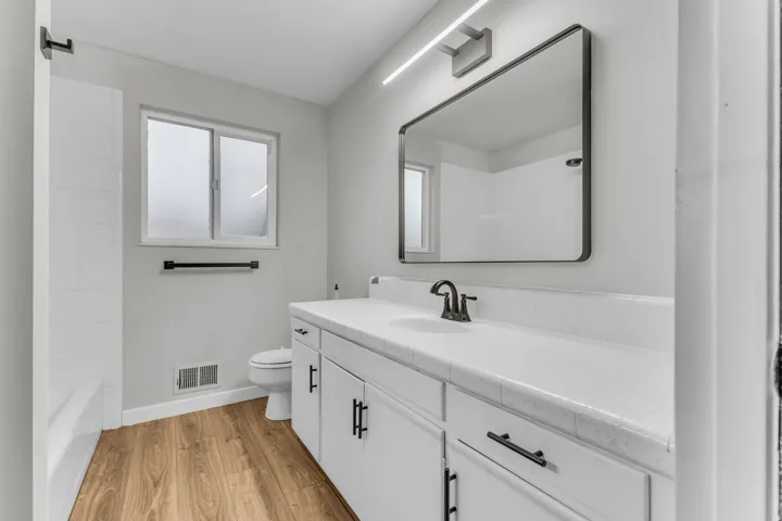Bathroom featuring a single vanity with white cabinetry, wood-finish flooring, a white bathtub with tile surround, and a rectangular mirror with integrated lighting